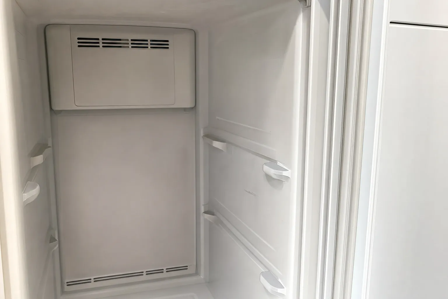 Tall empty compartment with a vent housing at the top of the rear wall and shelf supports along the door side